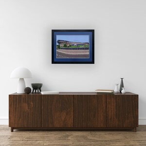 May include: A framed print of a stadium with a blue sky in the background. The stadium is made of concrete and has a large, curved roof. The print is in a black frame with a blue mat.