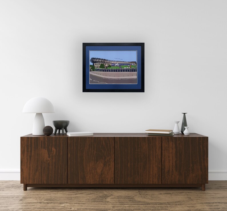May include: A framed print of a stadium with a blue sky in the background. The stadium is made of concrete and has a large, curved roof. The print is in a black frame with a blue mat.