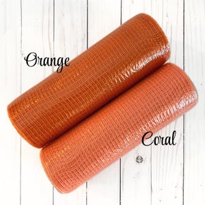 May include: Two rolls of decorative mesh in orange and coral colors. The orange roll is labeled "Orange" and the coral roll is labeled "Coral". The mesh has a woven texture and is wrapped in clear plastic. The background is a white wooden surface.