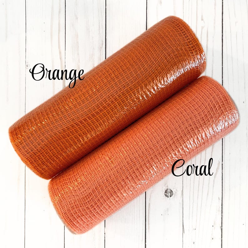 May include: Two rolls of decorative mesh in orange and coral colors. The orange roll is labeled "Orange" and the coral roll is labeled "Coral". The mesh has a woven texture and is wrapped in clear plastic. The background is a white wooden surface.