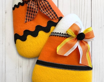 Puede incluir: Dos decoraciones de fieltro de caramelos de maíz. Cada una tiene una parte superior blanca, un centro naranja y una base amarilla, con ribete negro y un lazo decorativo. Los lazos presentan cintas naranjas, amarillas y blancas. Las decoraciones están sobre una superficie de madera blanca.