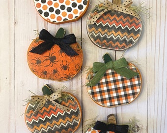May include: Six wooden pumpkins with different Halloween-themed patterns. The pumpkins are decorated with bows in black, green, and brown. The patterns include polka dots, zigzags, plaid, and spiders.