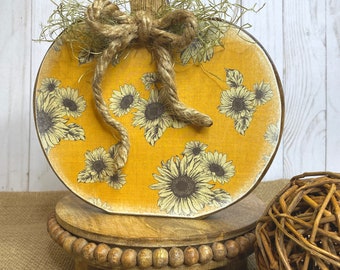 May include: A wooden pumpkin with a sunflower pattern and a burlap bow, sitting on a wooden pedestal with a beaded edge.