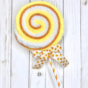 May include: A large, decorative lollipop with a yellow, white, and orange swirl design. The lollipop is adorned with a candy corn patterned bow and a striped orange and white stick. The background is a white wooden surface.
