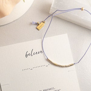 May include: A delicate gold bead bracelet with a lilac cord, presented with a card that says "believe" and "Believe in yourself, and you will be unstoppable." The card also features the brand name "lovassion."