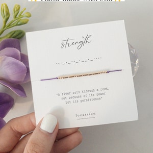 Peut inclure: Un bracelet violet et or avec le mot "strength" écrit en cursive sur une carte blanche. La carte comprend également la citation "A river cuts through a rock, not because of its power but its persistence." et le nom de la marque "lovassion".