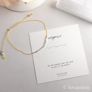 May include: A yellow string bracelet with silver-colored beads and a Morse code design. The bracelet is next to a card that says "inspire" and "Be the change that you wish to see in the world."