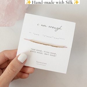 May include: A pink and gold bracelet with the words "i am enough" and "Good enough, strong enough, pretty enough, smart enough" on a white card. The bracelet is made with silk thread and gold beads. The card also has the word "lovassion" on it.