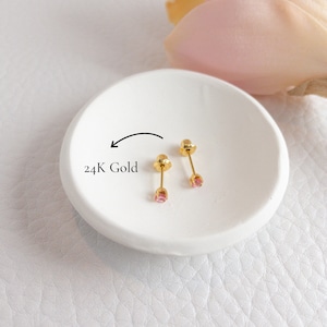 May include: Two small, gold-coloured stud earrings with pink gemstones are displayed on a white, circular dish. The text "24K Gold" is next to the earrings. The background is a soft, blurred arrangement of white and pink.