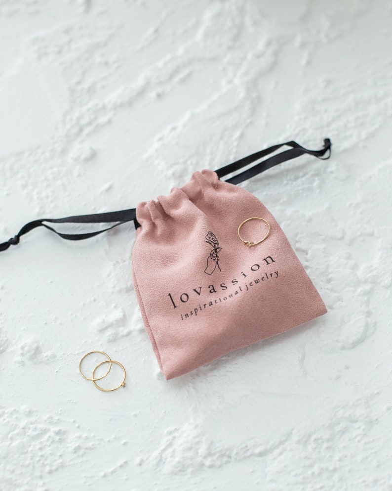 May include: A pink velvet pouch with a black drawstring and the text "lovassion inspirational jewellery". Two gold rings are visible outside the pouch.