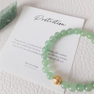 May include: A green aventurine bead bracelet with a gold-colored accent bead. A card with the word "Protection" and text about aventurine's protective properties is next to the bracelet. The brand name "lovassion" is visible on the card.