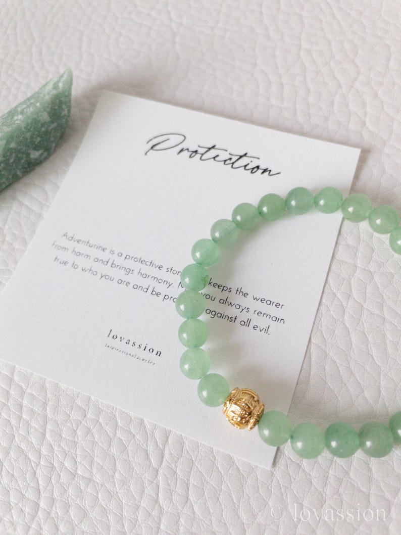 May include: A green aventurine bead bracelet with a gold-colored accent bead. A card with the word "Protection" and text about aventurine's protective properties is next to the bracelet. The brand name "lovassion" is visible on the card.