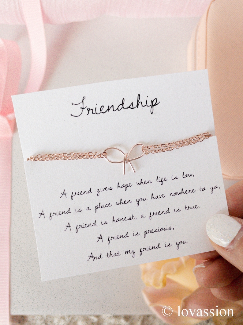 May include: A white card with the word "Friendship" written in cursive at the top. The card has a rose gold chain necklace with a bow charm on it. The card also has a handwritten message that reads: "A friend gives hope when life is low, A friend is a place when you have nowhere to go, A friend is honest, a friend is true. A friend is precious, And that my friend is you."