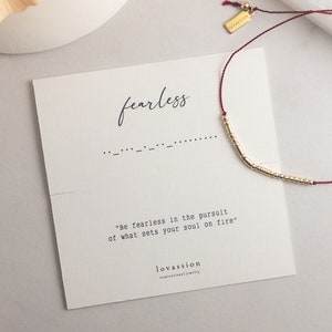 Peut inclure: Un bracelet à perles dorées avec un cordon rouge est présenté à côté d'une carte qui dit "fearless". La carte comprend également la citation : "Soyez sans crainte dans la poursuite de ce qui enflamme votre âme."