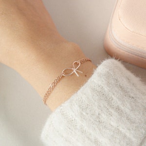May include: A delicate rose gold chain bracelet with a small bow charm. The bracelet is on a person's wrist.