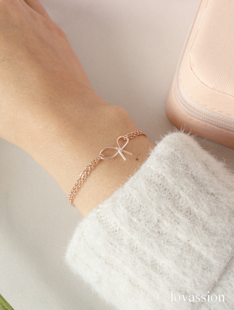 May include: A delicate rose gold chain bracelet with a small bow charm. The bracelet is on a person's wrist.