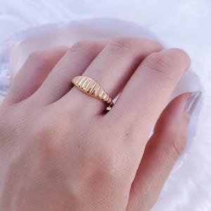 May include: A gold ring with a ribbed design. The ring is worn on a finger. The ring's design features a series of parallel, curved ridges, creating a textured surface. The ring is a simple yet elegant piece of jewellery.