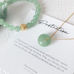 May include: A light green aventurine bracelet with a gold accent bead and a matching pendant necklace. The pendant is a circular jade stone on a gold chain. A card reads "Protection" and describes the stone's properties.