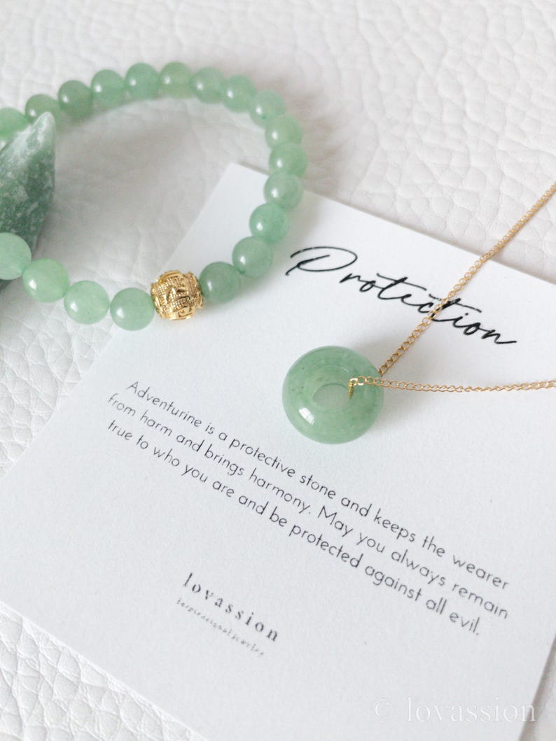 May include: A light green aventurine bracelet with a gold accent bead and a matching pendant necklace. The pendant is a circular jade stone on a gold chain. A card reads "Protection" and describes the stone's properties.