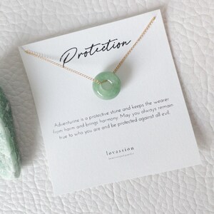 May include: A delicate gold chain necklace featuring a round, light green aventurine pendant. The pendant is centered on a white card with the word "Protection" and text about the stone's properties. The card is on a white textured surface.