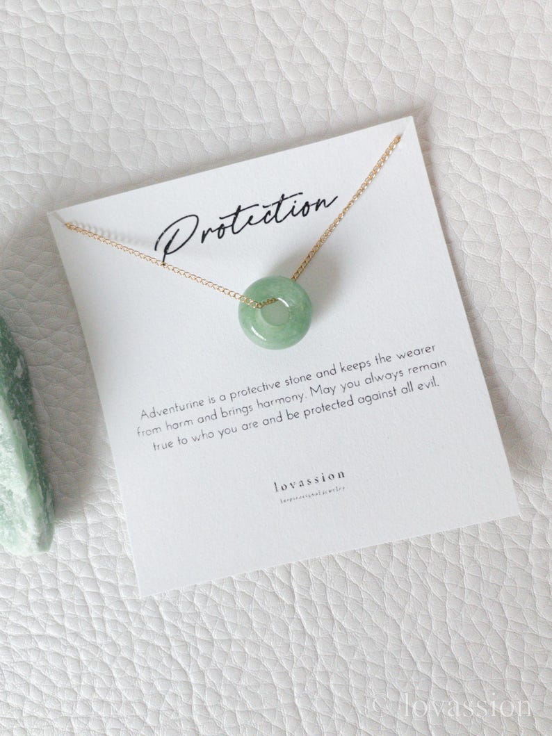 May include: A delicate gold chain necklace featuring a round, light green aventurine pendant. The pendant is centered on a white card with the word "Protection" and text about the stone's properties. The card is on a white textured surface.