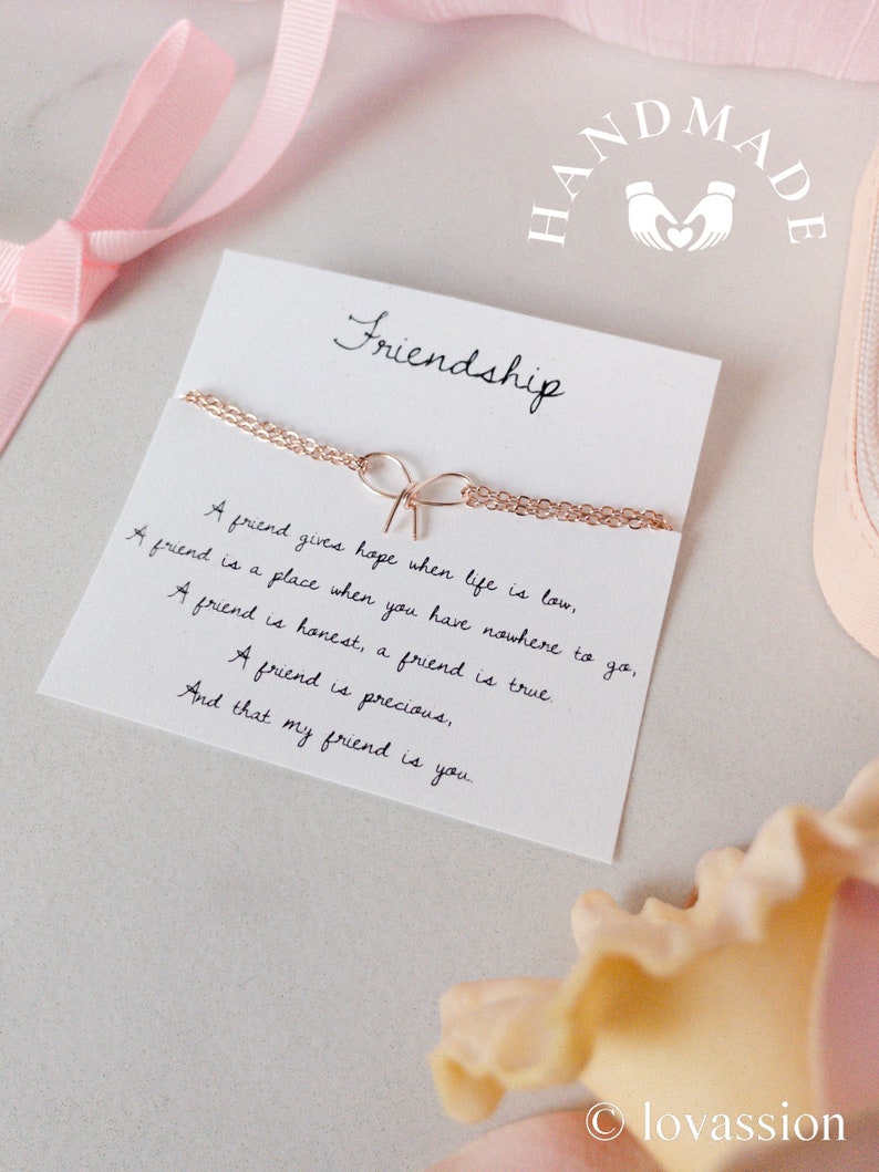 May include: A white card with the word "Friendship" written in black cursive. The card features a gold chain necklace with a bow charm. The card also has a quote about friendship written in black cursive. The card is on a white surface with a pink ribbon and a yellow flower.