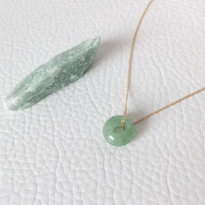 May include: A light green jade donut pendant necklace with a gold chain. A raw, light green jade stone is next to the necklace. The necklace and stone are on a white, textured surface.