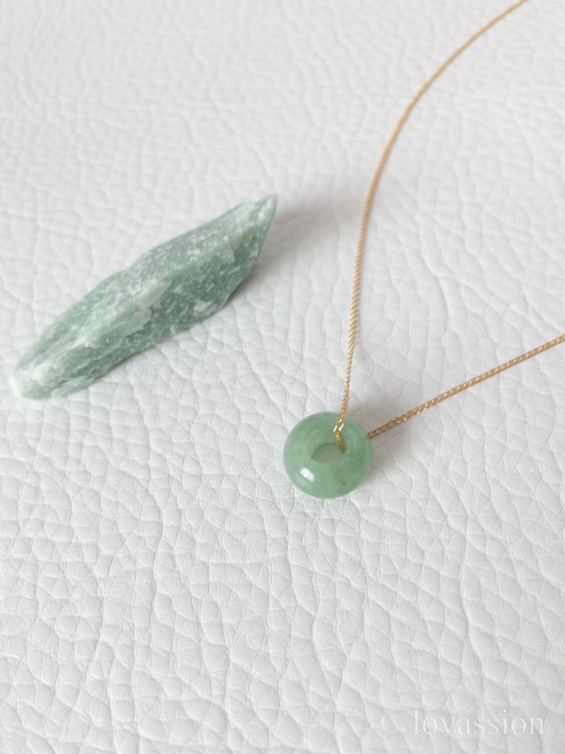 May include: A light green jade donut pendant necklace with a gold chain. A raw, light green jade stone is next to the necklace. The necklace and stone are on a white, textured surface.