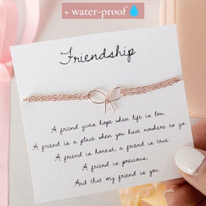 May include: A white card with the word "Friendship" written in black cursive. The card features a quote about friendship and a delicate gold chain necklace with a bow charm. The card is tarnish-resistant and water-proof.