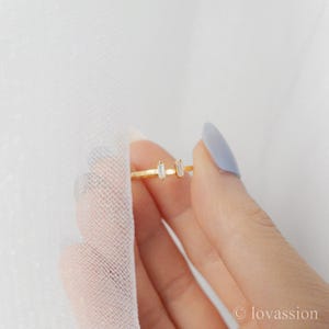 May include: A delicate gold ring with two rectangular, clear gemstones. The ring is held by a hand with a light blue painted nail. The background is a soft, white fabric. The ring is a minimalist jewelry piece.