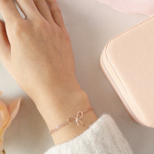 May include: A delicate rose gold chain bracelet with a small bow charm. The bracelet is on a person's wrist.