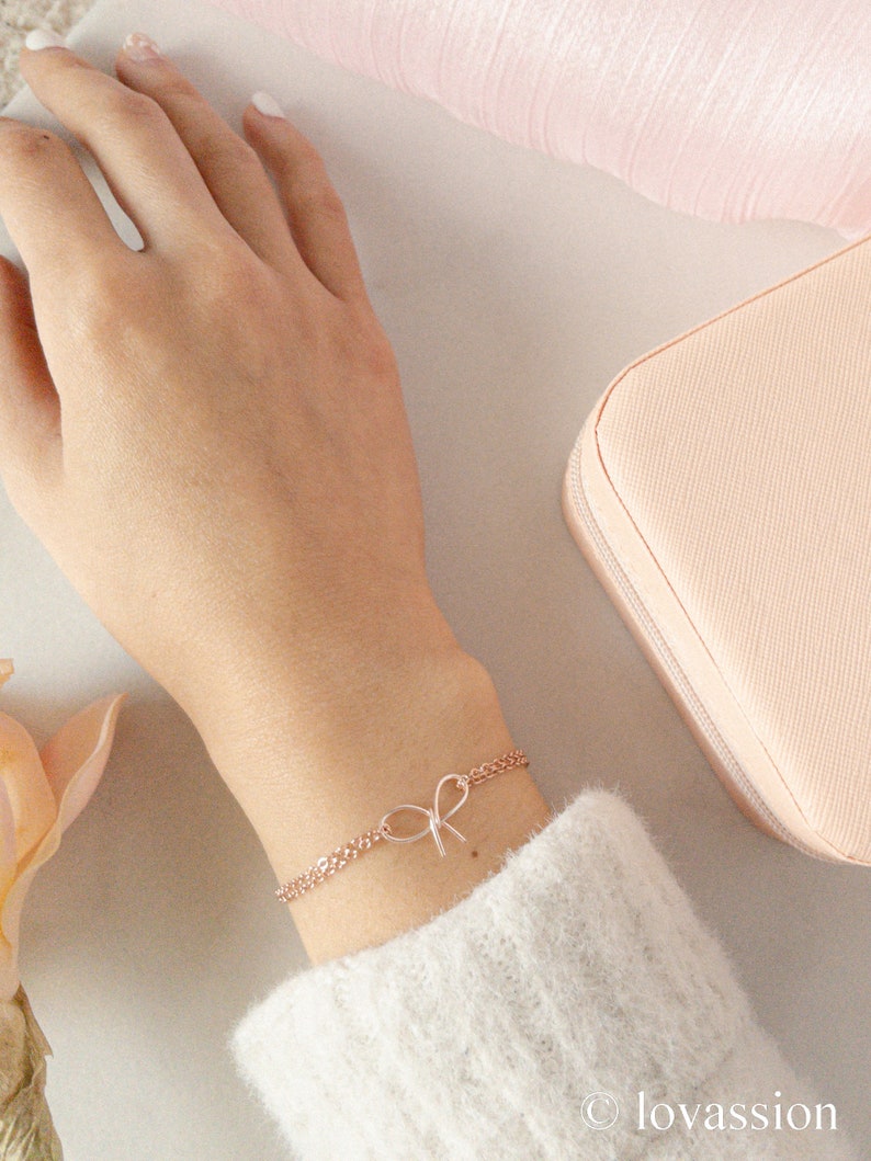May include: A delicate rose gold chain bracelet with a small bow charm. The bracelet is on a person's wrist.