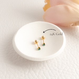 May include: A pair of gold stud earrings with teal-colored gemstones, displayed on a white, round dish. The earrings are labeled as 24K gold. The background is a soft, neutral color.