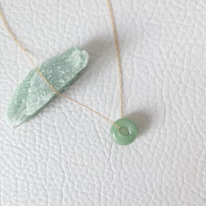 May include: A delicate gold necklace featuring a small, round, light green jade pendant. The pendant is a smooth, polished circle. The necklace is displayed on a white, textured surface, with a piece of raw green jade nearby.