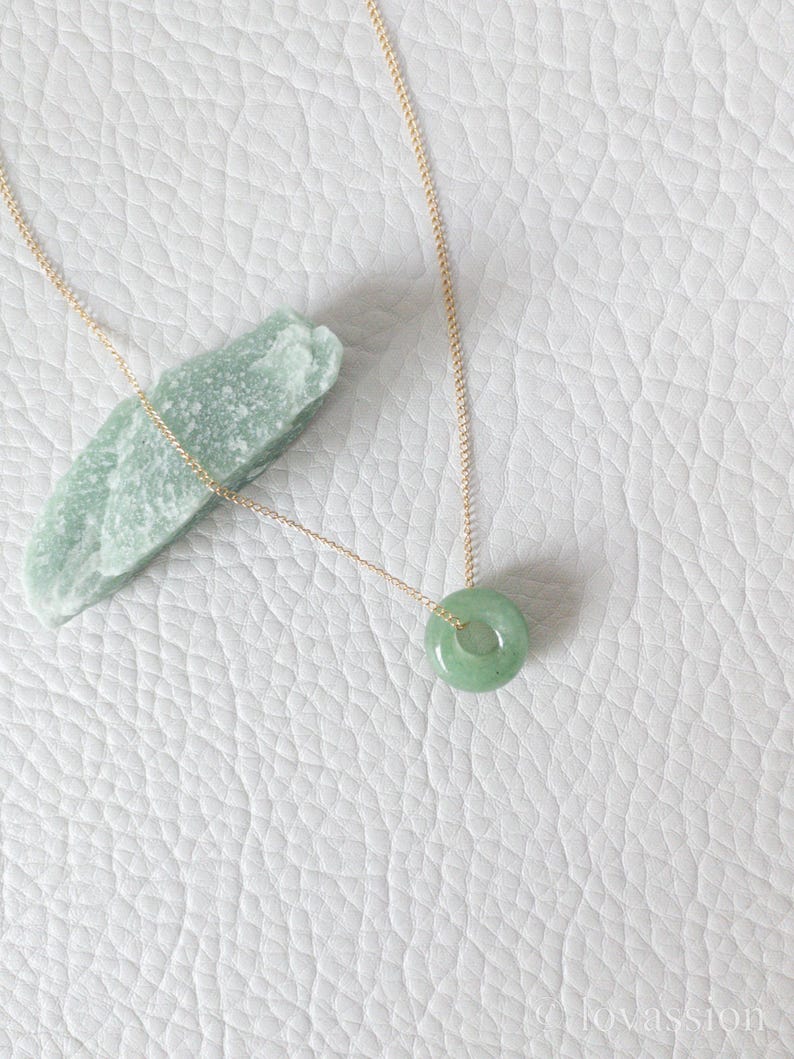 May include: A delicate gold necklace featuring a small, round, light green jade pendant. The pendant is a smooth, polished circle. The necklace is displayed on a white, textured surface, with a piece of raw green jade nearby.