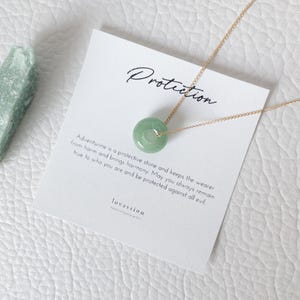 May include: A gold chain necklace with a circular, light green aventurine pendant. The pendant is centered on a white card with the word "Protection" and text about aventurine. A piece of green stone is on the left.