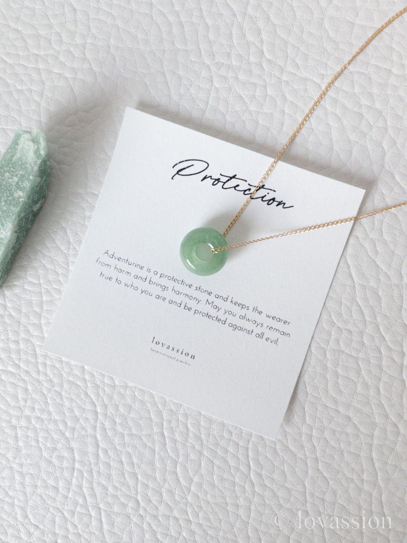 May include: A gold chain necklace with a circular, light green aventurine pendant. The pendant is centered on a white card with the word "Protection" and text about aventurine. A piece of green stone is on the left.