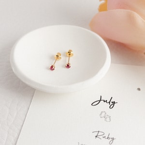 May include: A pair of gold stud earrings with small, round, red gemstones. The earrings are displayed in a white, shallow dish. A card with the word "July" and "Ruby" is visible.