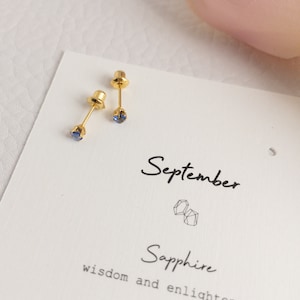 May include: A pair of gold-colored stud earrings with small, round, blue gemstones. The earrings are displayed on a white card with the word "September" and "Sapphire" printed on it. The card also includes the phrase "wisdom and enlightenment".