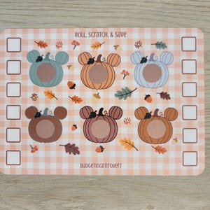 May include: A rectangular, fall-themed scratch-off game board with six pumpkin designs in various colors. The board features the text "ROLL, SCRATCH, & SAVE" and is decorated with leaves and acorns. The background has a peach and white gingham pattern.