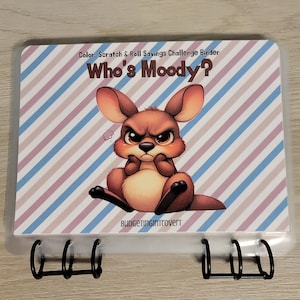 May include: A clear plastic binder with a colorful cover featuring a cartoon kangaroo with an angry expression. The cover has the text "Color, Scratch & Roll Savings Challenge Binder" and "Who's Moody?" The bottom of the cover has the text "budgetingintrovert".