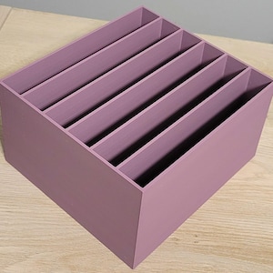 May include: A mauve-colored wooden organizer with six angled compartments. The open-top box is designed to hold and sort items. The organizer is made of wood and has a smooth, painted finish. The box sits on a light-colored wooden surface.