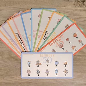 May include: A set of colorful flashcards with months of the year printed on them. Each card has a different pastel border. The January card has illustrations of a snowman, a mug, and a boot. The cards are arranged in a fan shape.