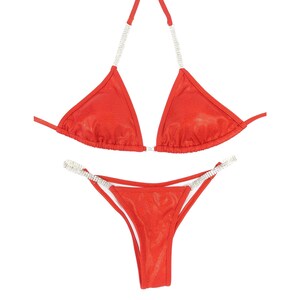 May include: Red bikini set with rhinestone accents. The triangle top has rhinestone straps, and the bottoms have rhinestone detailing along the sides. The fabric has a subtle shimmer.