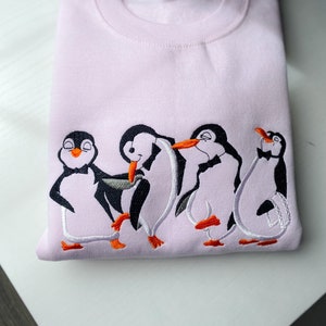 May include: A pink sweatshirt with an embroidered design of four penguins wearing bowties. The penguins are in a line, with the first two penguins facing each other and the last two penguins facing forward. The penguins are embroidered in white, black, and orange.