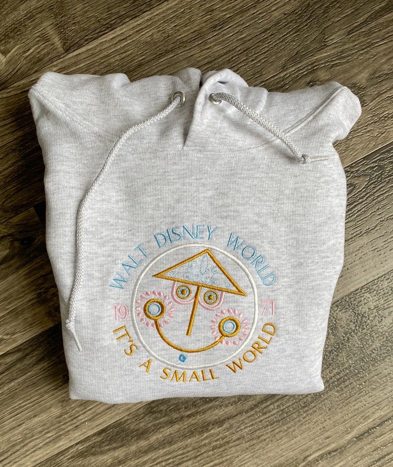 May include: Light gray hoodie with a drawstring hood. The front features an embroidered design with the text "Walt Disney World" and "It's a Small World" in a circular pattern, along with a smiling face graphic and the number "1971".