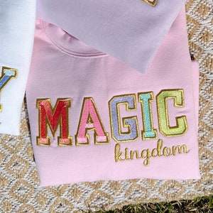 May include: Pink sweatshirt with the word "MAGIC" embroidered in rainbow colors with a gold outline. The word "kingdom" is embroidered in gold below the word "MAGIC".