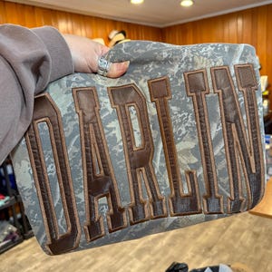 May include: A camouflage-patterned bag with the word "DARLIN" embroidered in brown block letters. The bag has a rectangular shape and appears to be made of a soft, textured fabric. The background has a camouflage pattern in shades of green, brown, and gray.