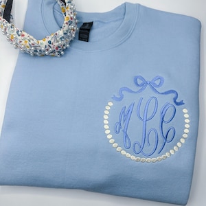 May include: A light blue sweatshirt with a white pearl-like circle around a blue monogram with a blue bow on top.