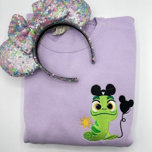 May include: A folded lavender sweatshirt with an embroidered green chameleon character holding a sun and a Mickey Mouse balloon. A sequined bow headband with a black band rests on top.